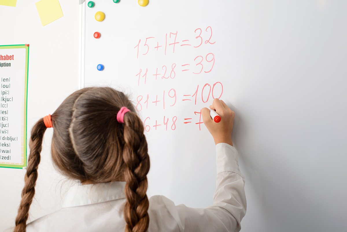 Education, elementary school, learning, math and people concept. Clever female learner of primary school writing on board some mathematical equations, counting numbers